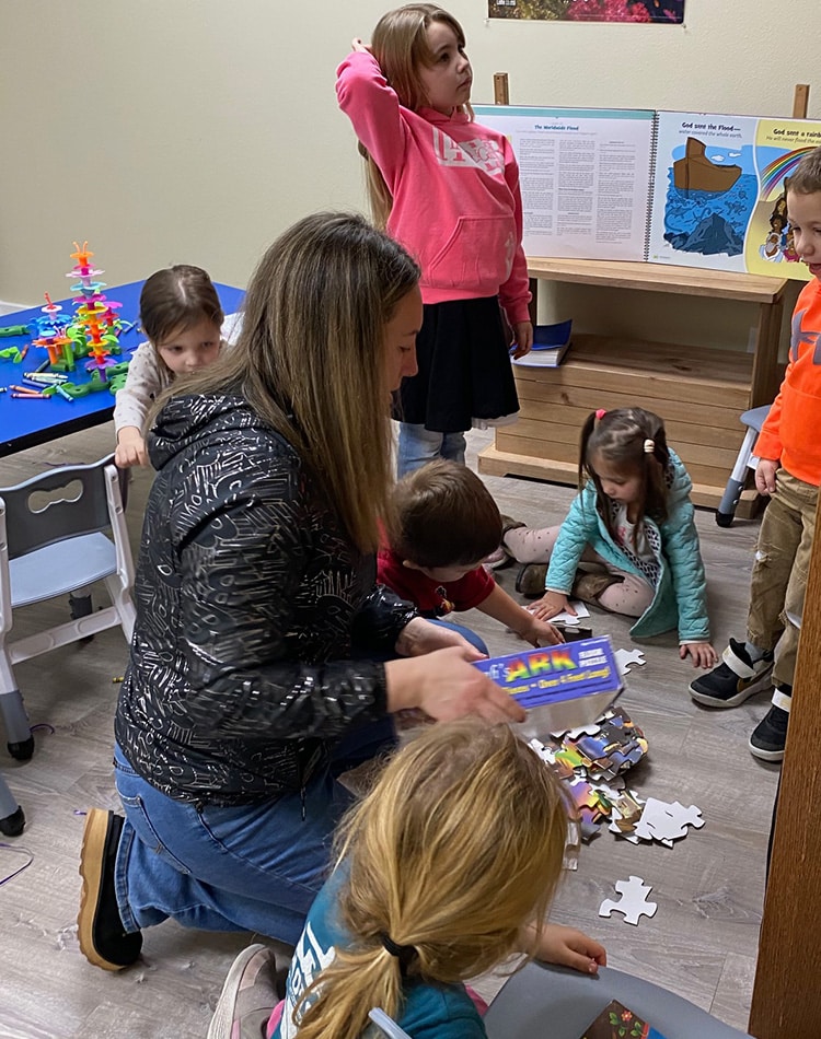 An adult and several young children gather on the floor and interact with puzzle pieces in a classroom setting.