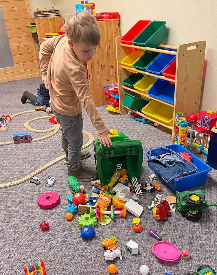 A young boy stands next to a tipped-over green bin with toys scattered on the carpet in a playroom. Another child is playing in the background.