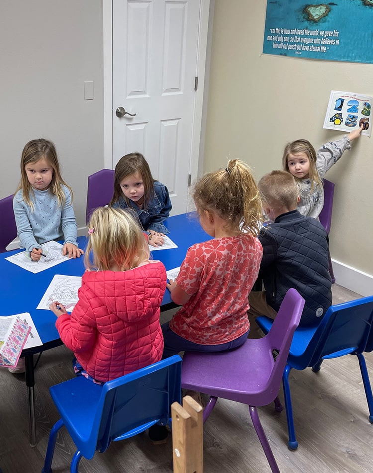 Seven young children sit around a blue table coloring on paper in a classroom with purple chairs and educational posters on the walls.